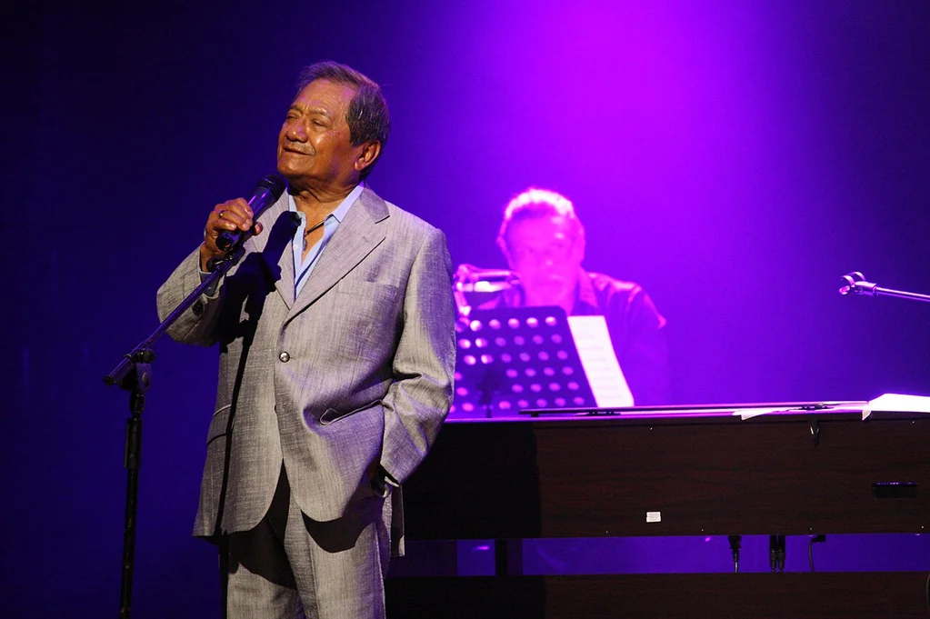 Armando Manzanero cantando en un escenario morado, junto a un teclado