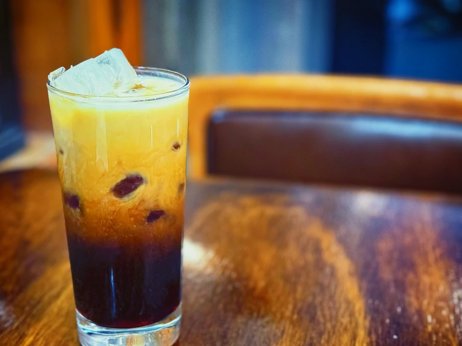 Vaso alto con café helado y leche en capas sobre hielo, servido en una mesa de madera