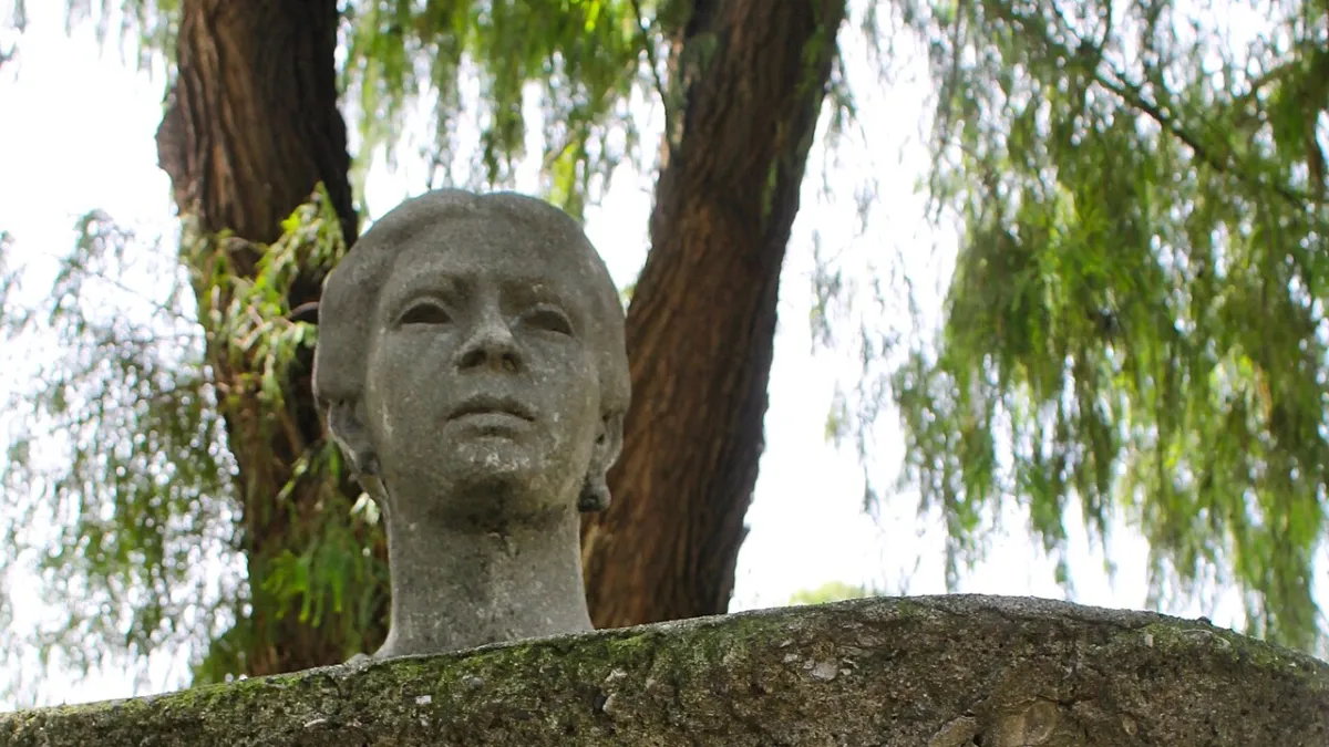 Busto gris de Dolores Bedoya de Molina sobre una base de piedra, con árboles al fondo