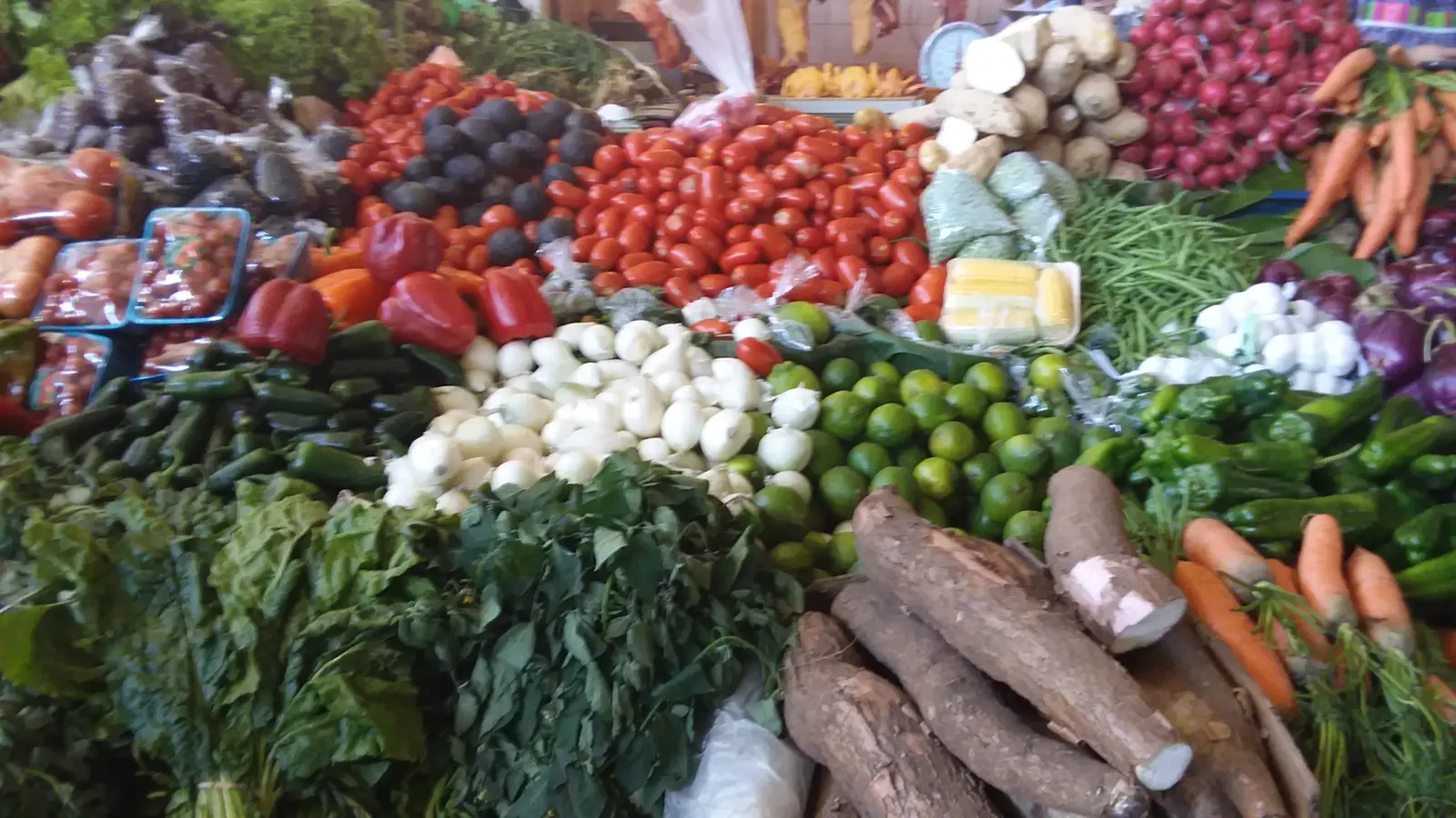 Puesto de mercado con montones de tomate, cebolla, limón, güisquil, yuca, zanahoria y hojas verdes