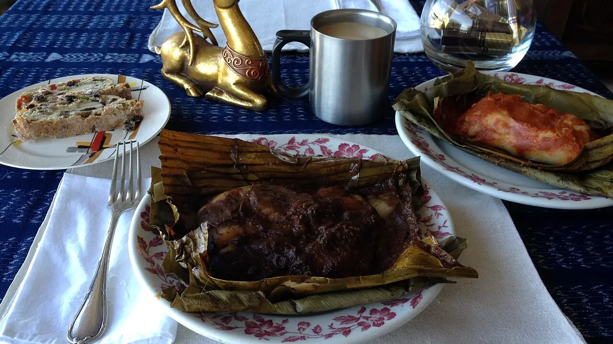 Tamal envuelto en hoja de plátano servido en un plato floreado, junto a otro tamal de masa roja y una taza de café