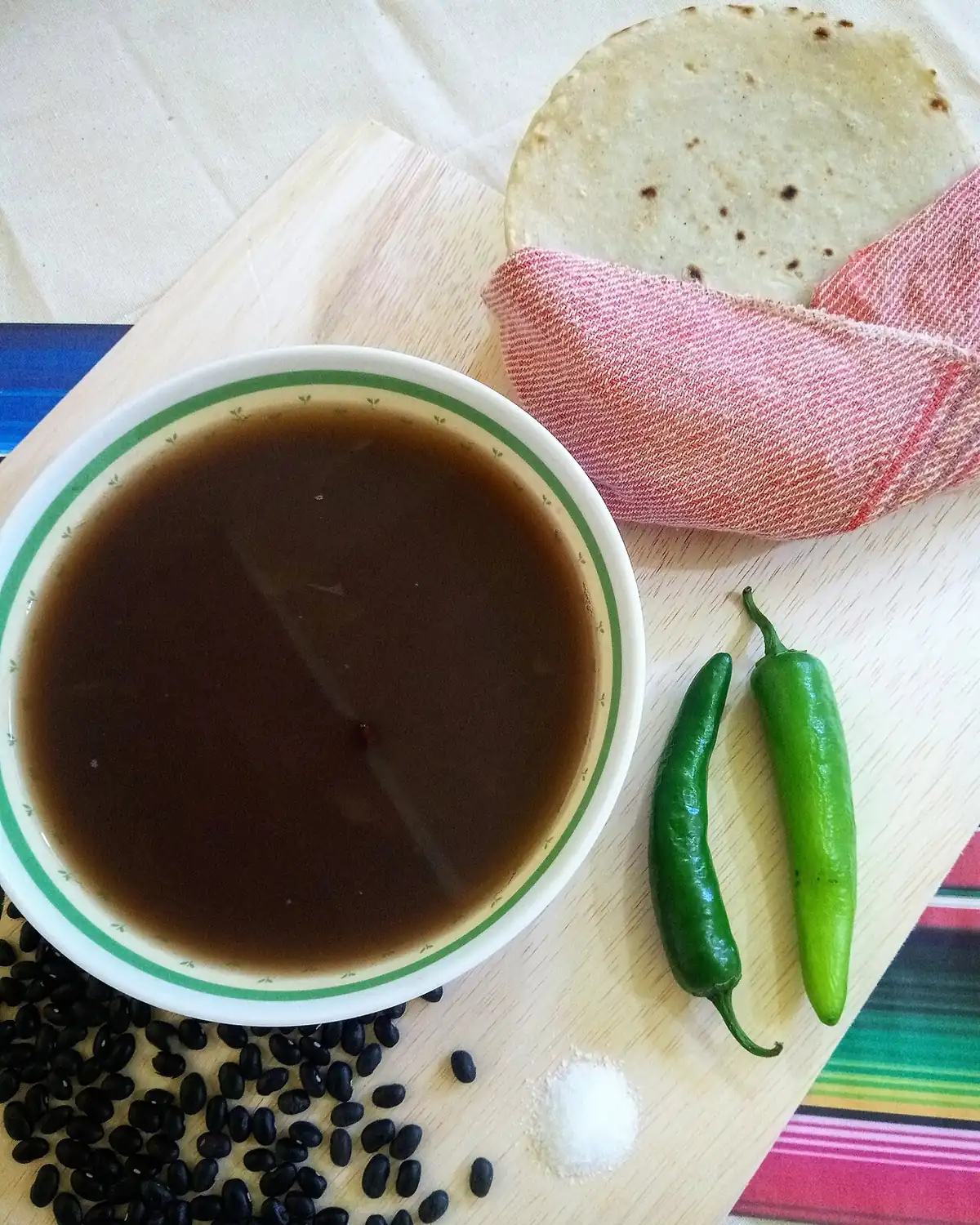 Tazón de caldo oscuro de frijol negro con una tortilla, chiles verdes y frijoles secos sobre una tabla de madera