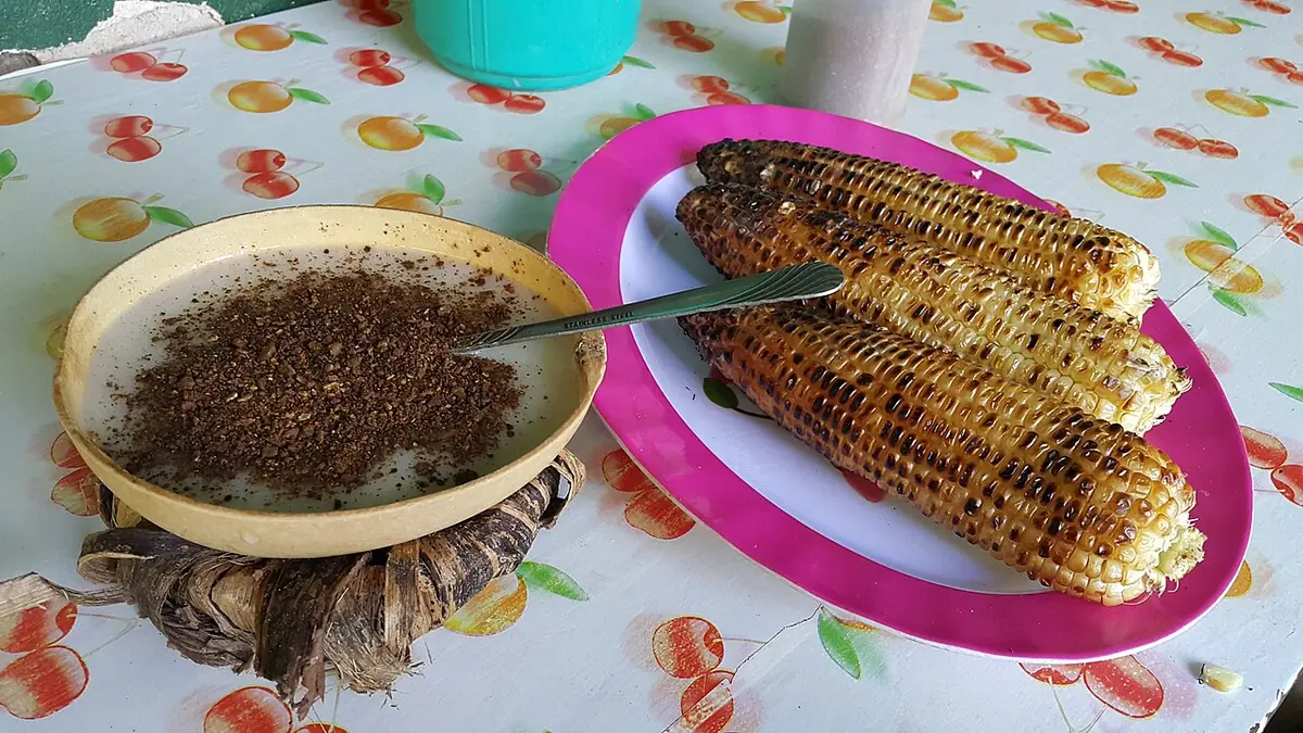 Mazorcas de maíz asadas en un plato rosado, junto a un tazón de barro con un polvo oscuro molido sobre una mesa