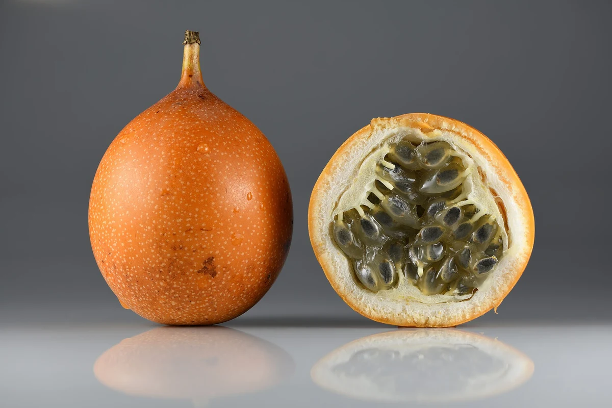 Una granadilla entera de cáscara anaranjada con pecas y otra partida a la mitad que muestra el anillo de cáscara, la pulpa blanca y las semillas negras envueltas en jugo