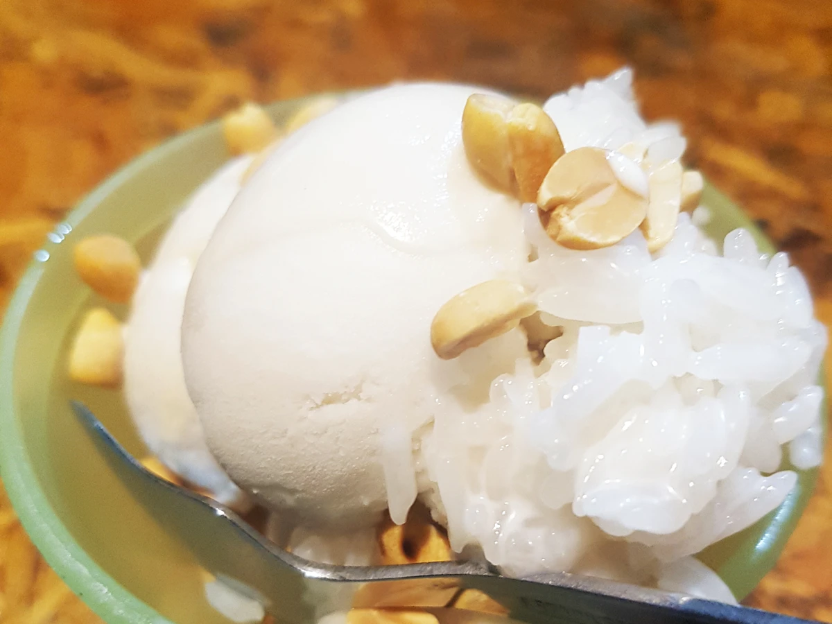 Bola de helado de coco blanco en un tazón verde, cubierta con coco natural y maní tostado, con una cuchara al lado