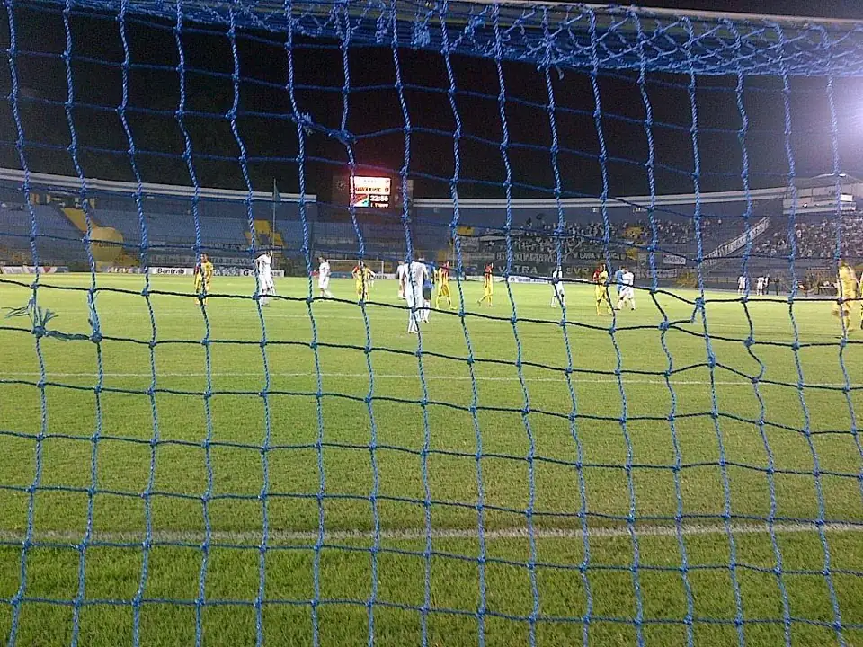 Partido nocturno visto a través de la red de una portería, con la gradería sur y el marcador al fondo