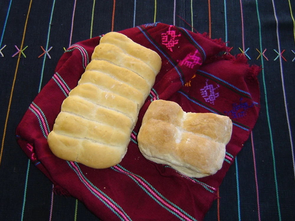 Dos piezas de pan francés guatemalteco, una alargada con cortes en la superficie y otra redonda, sobre un tejido rojo con franjas de colores