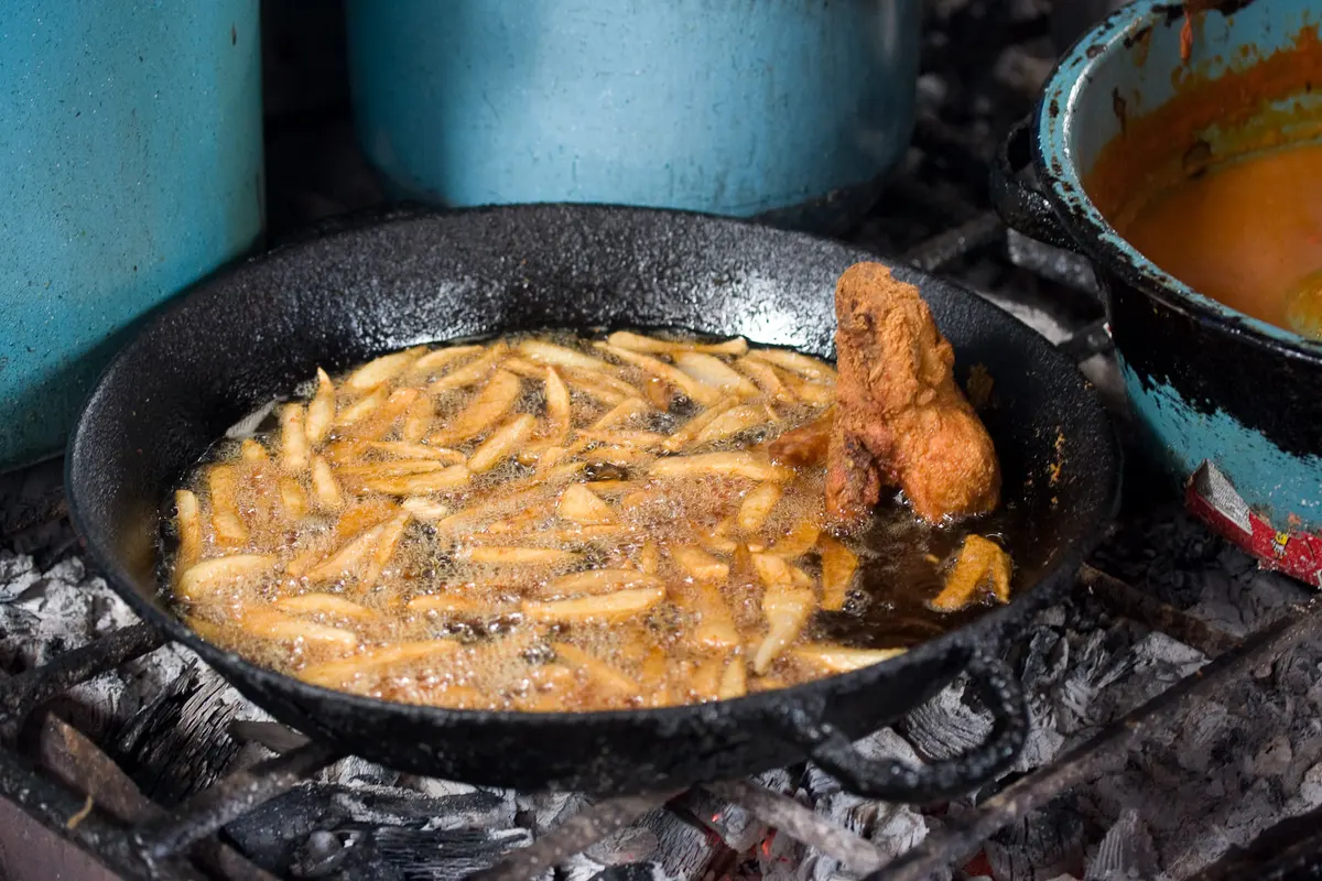 Bastones de papa friendo en un comal de hierro sobre carbón, junto a una pieza de pollo frito, en un puesto de mercado