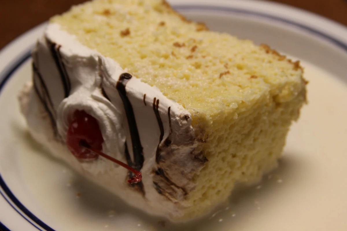 Porción de pastel tres leches con crema batida, hilo de chocolate y una cereza, servida en un plato blanco con borde azul