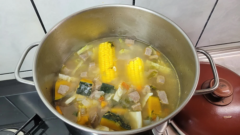 Olla de metal con caldo de carne, mazorcas de maíz y verduras en trozos grandes, recién retirada del fuego