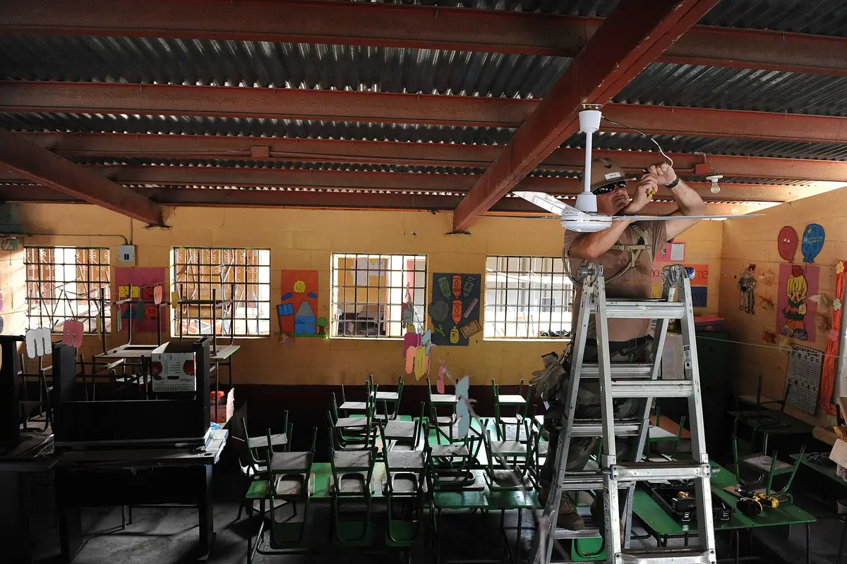 Un trabajador sobre una escalera instala un ventilador de techo en un aula con pupitres verdes