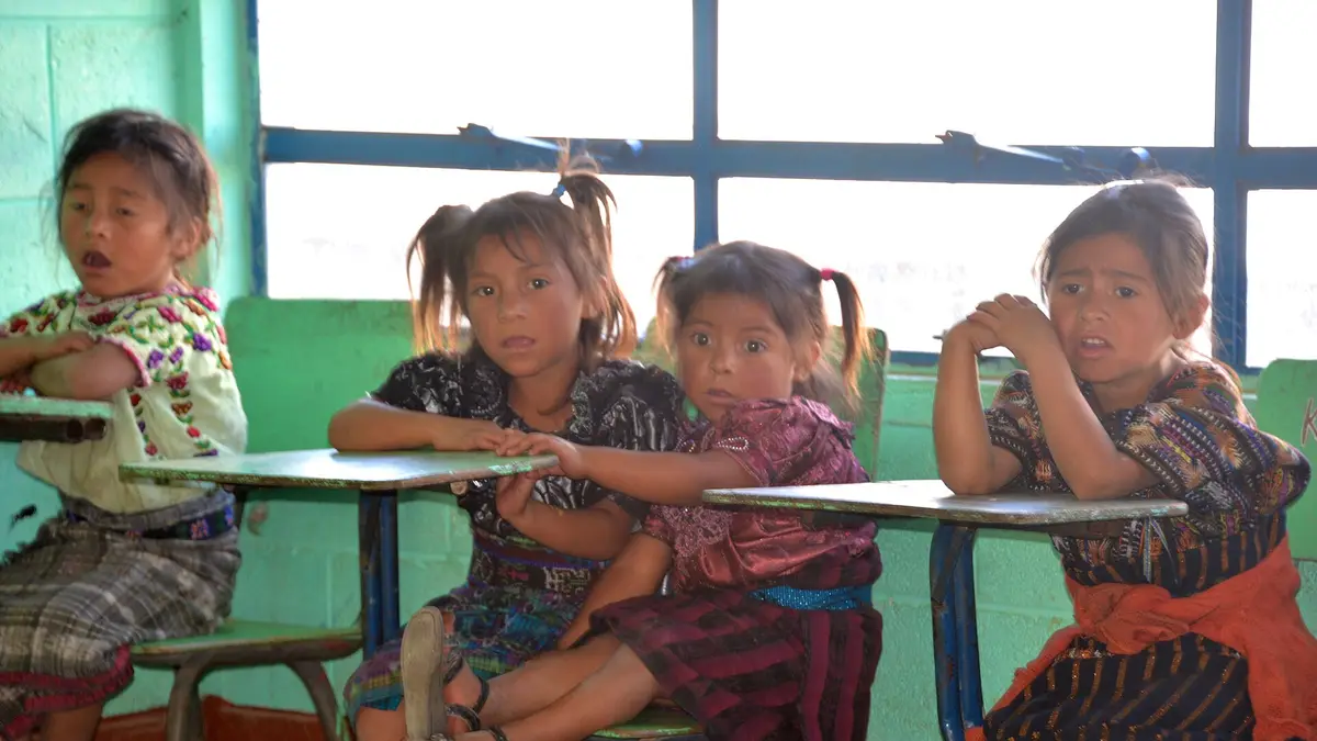 Cuatro niñas con traje típico sentadas en pupitres frente a una ventana en un aula rural de Guatemala