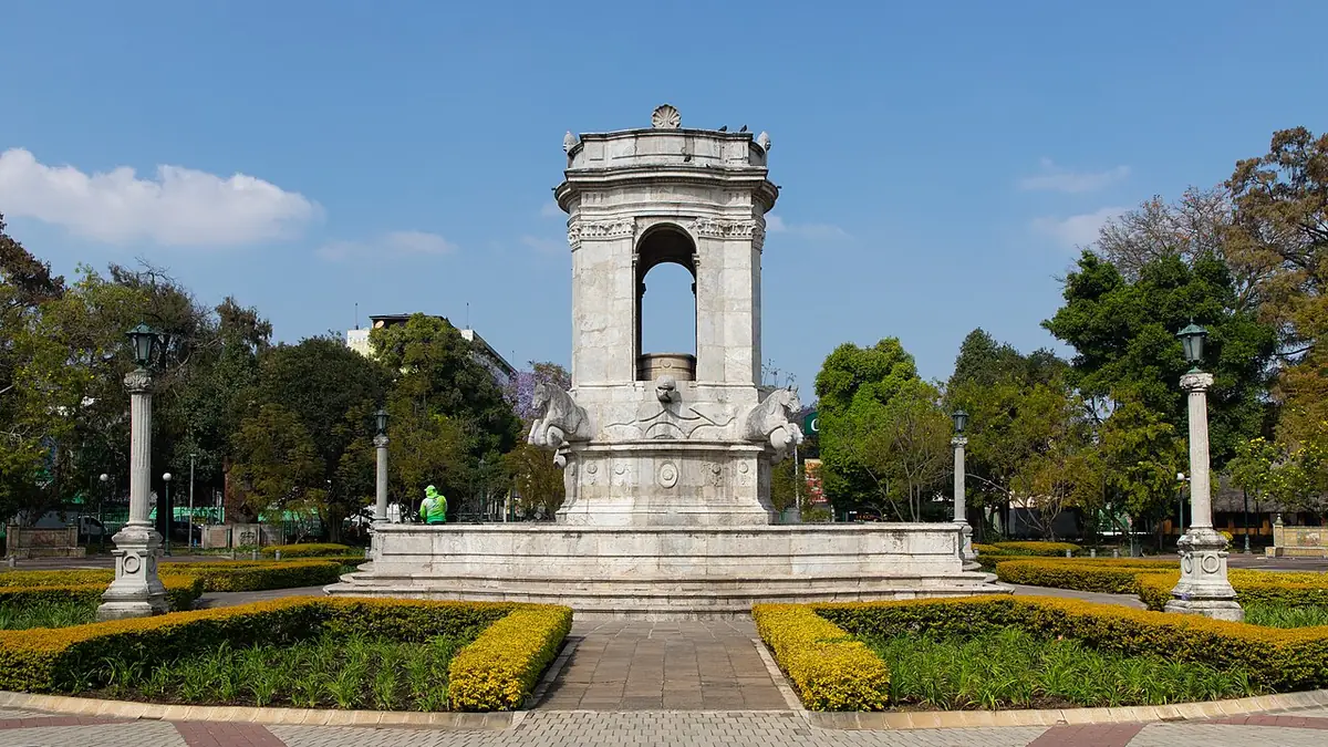 Plaza España en la Ciudad de Guatemala, con una fuente de piedra blanca, jardines y árboles bajo cielo azul