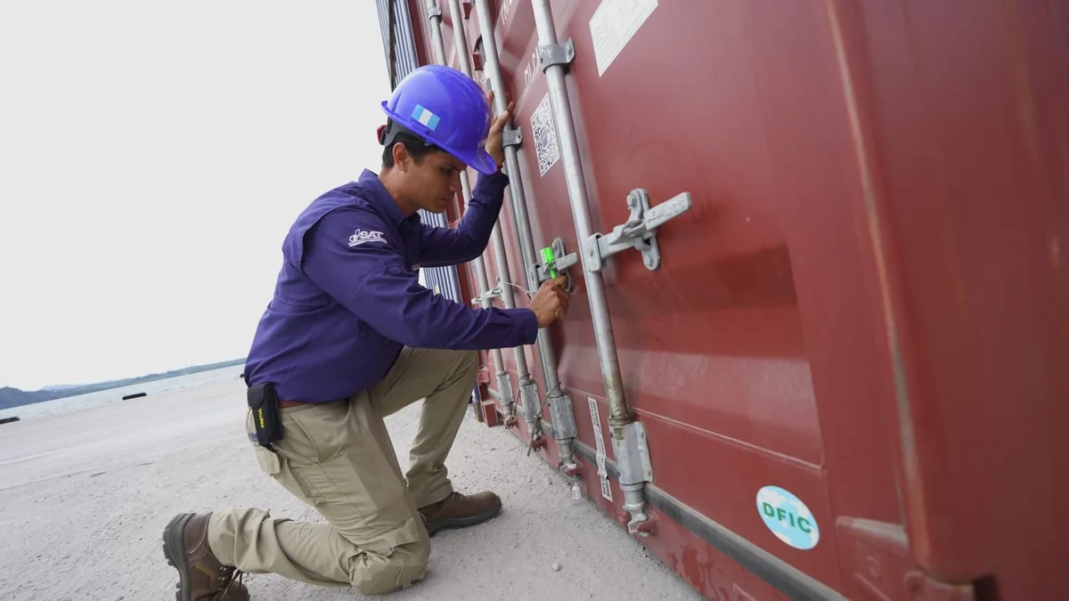 Técnico de la SAT con casco azul y uniforme morado cierra el seguro de un contenedor de carga rojo