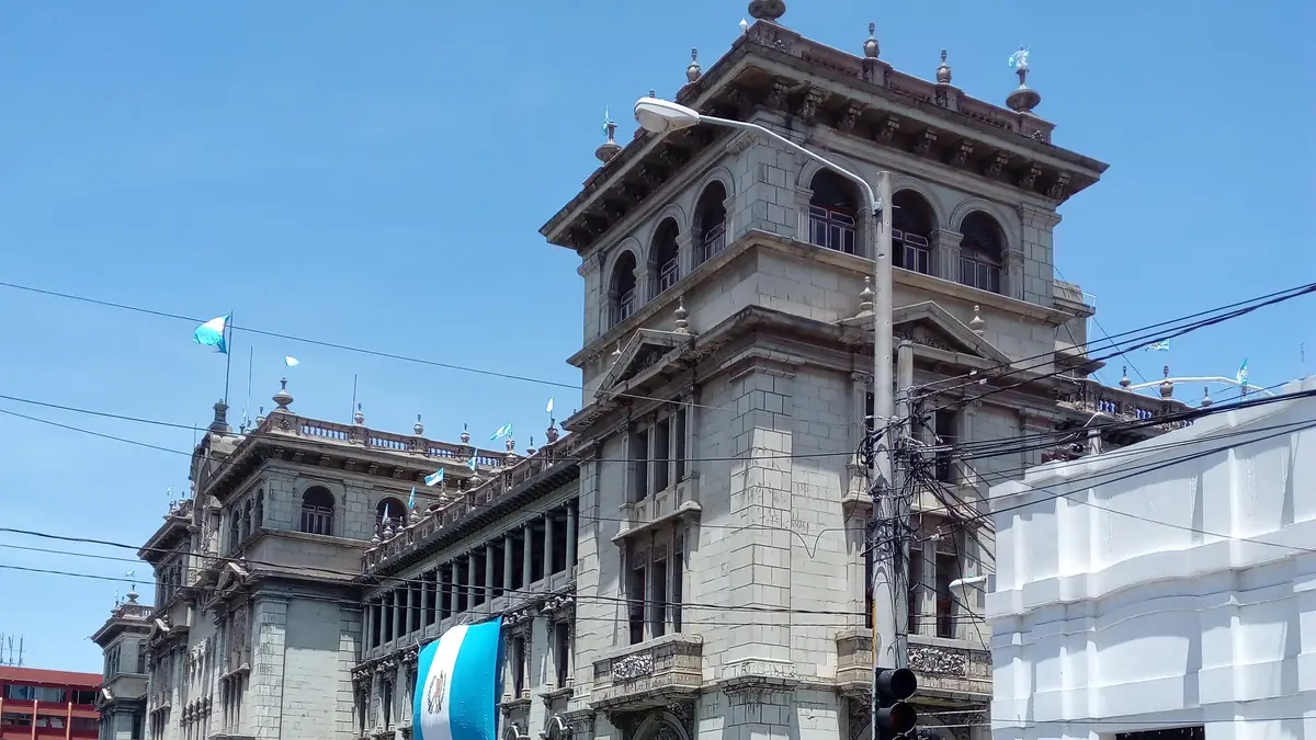Fachada lateral del Palacio Nacional de Guatemala, con piedra gris, bandera nacional y tránsito en la calle