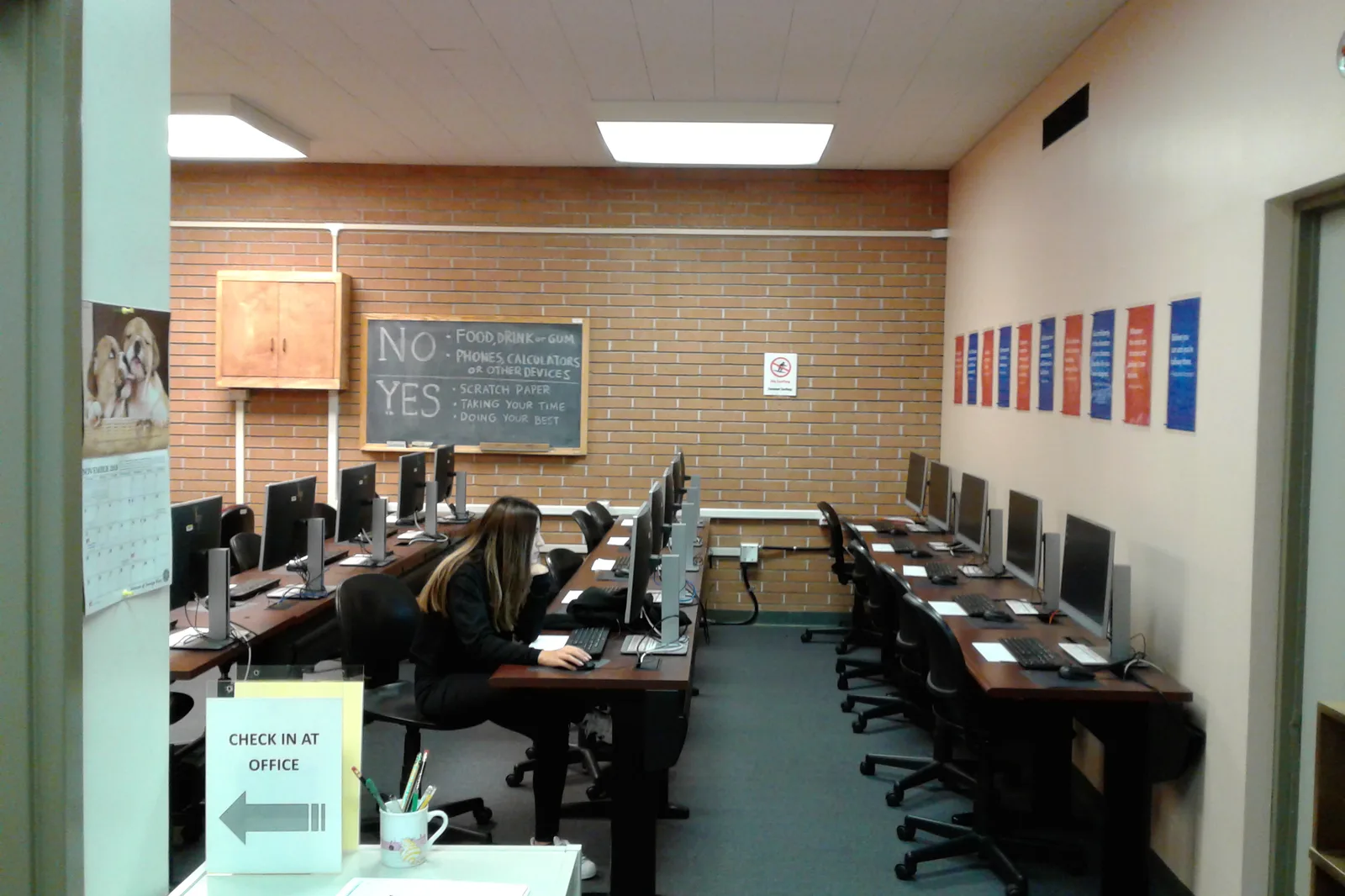 Sala de exámenes con varias computadoras alineadas, una persona respondiendo en una de las estaciones y las reglas de la prueba escritas en la pizarra